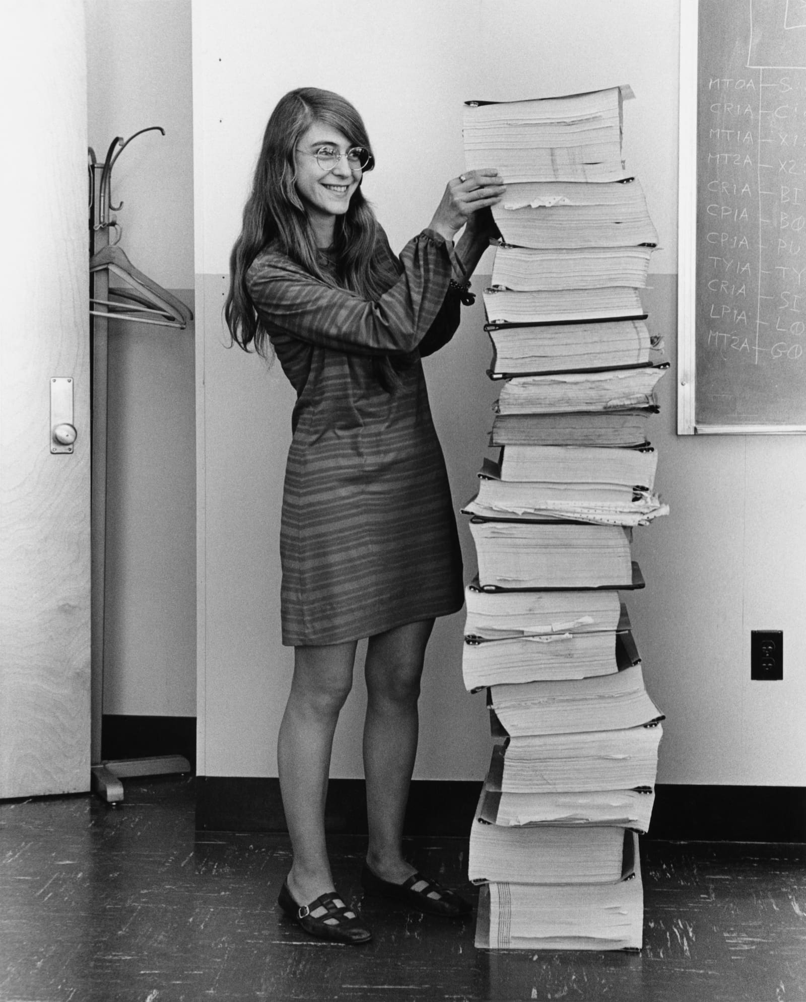 Black and white photograph: Margaret Hamilton standing next to a stack of printed program listings as tall as she is.
