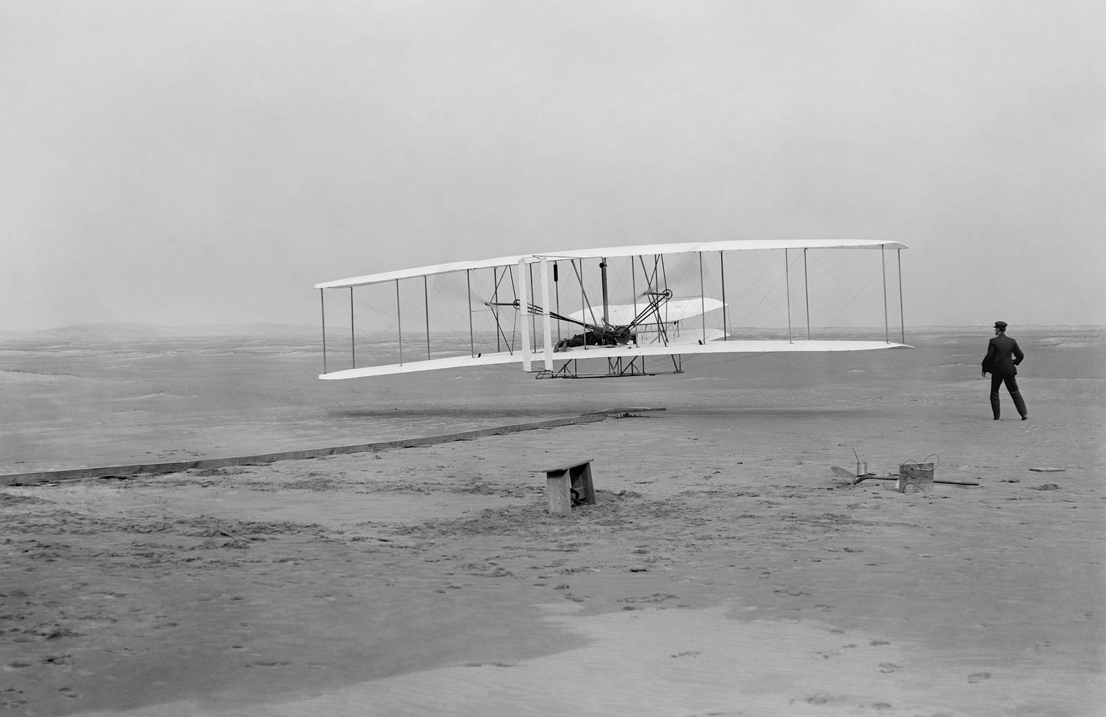 Black and white photograph: the Wright Flyer a few feet above the sand, Orville lying at the controls, Wilbur standing to the right.
