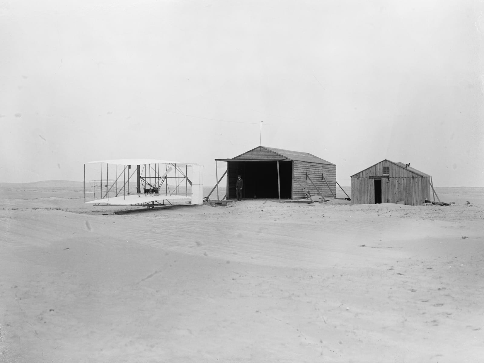Photographie en noir et blanc : le Flyer de 1903 sur son rail de lancement devant deux bâtiments de bois, sur le sable.
