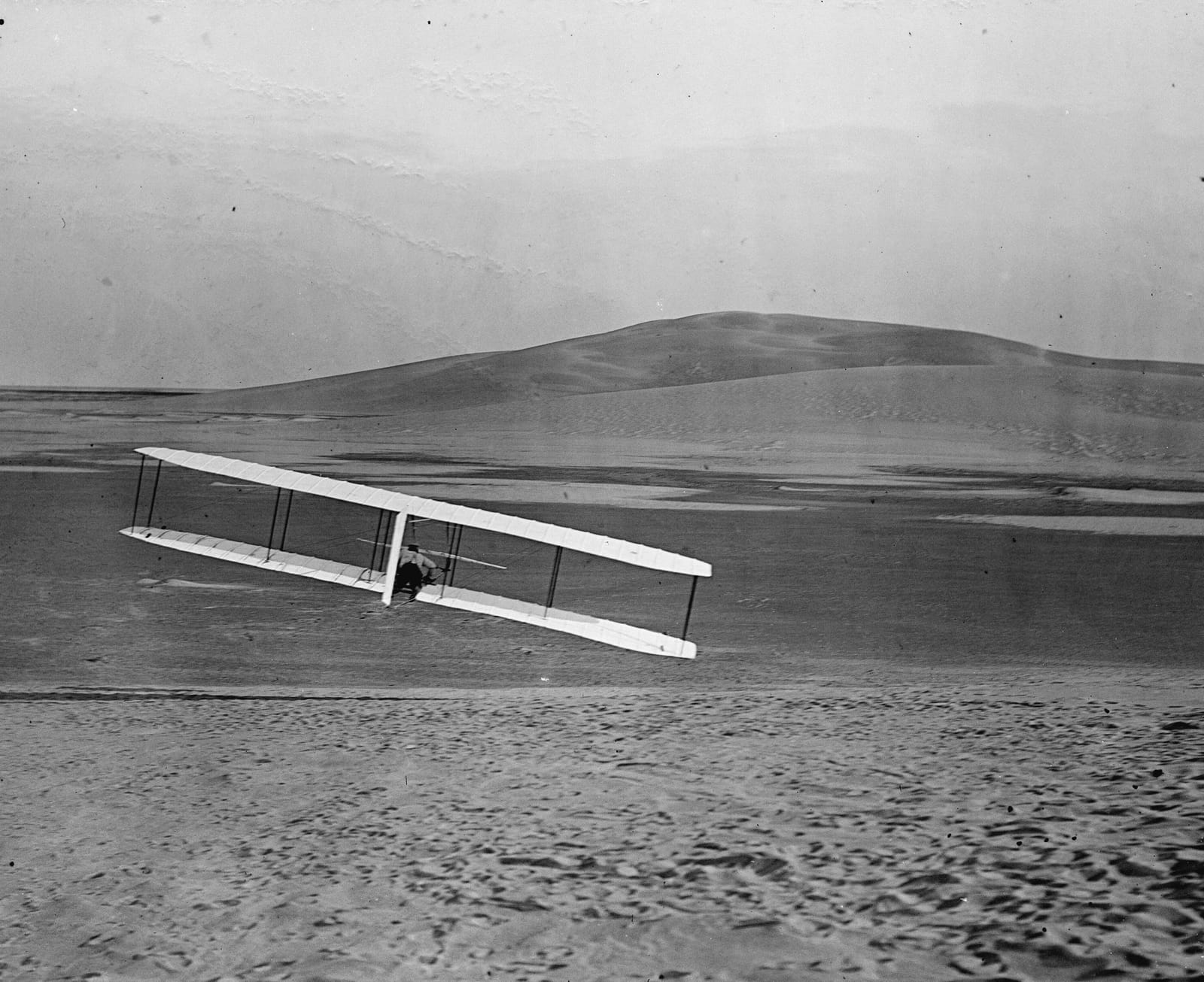 Black and white photograph: a biplane glider banking over a sand dune, seen from behind.